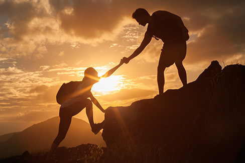 Photo of one person helping another up a mountain with a sunset in the background.