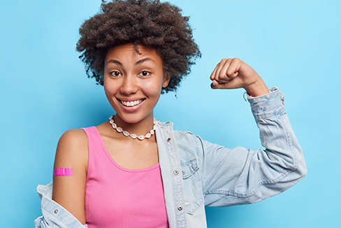 Photo of satisfied curly haired woman raises arm shows biceps wears pink t shirt denim shirt necklace adhesive bandage on shoulder after vaccination feels good and protected encourages to vaccinate